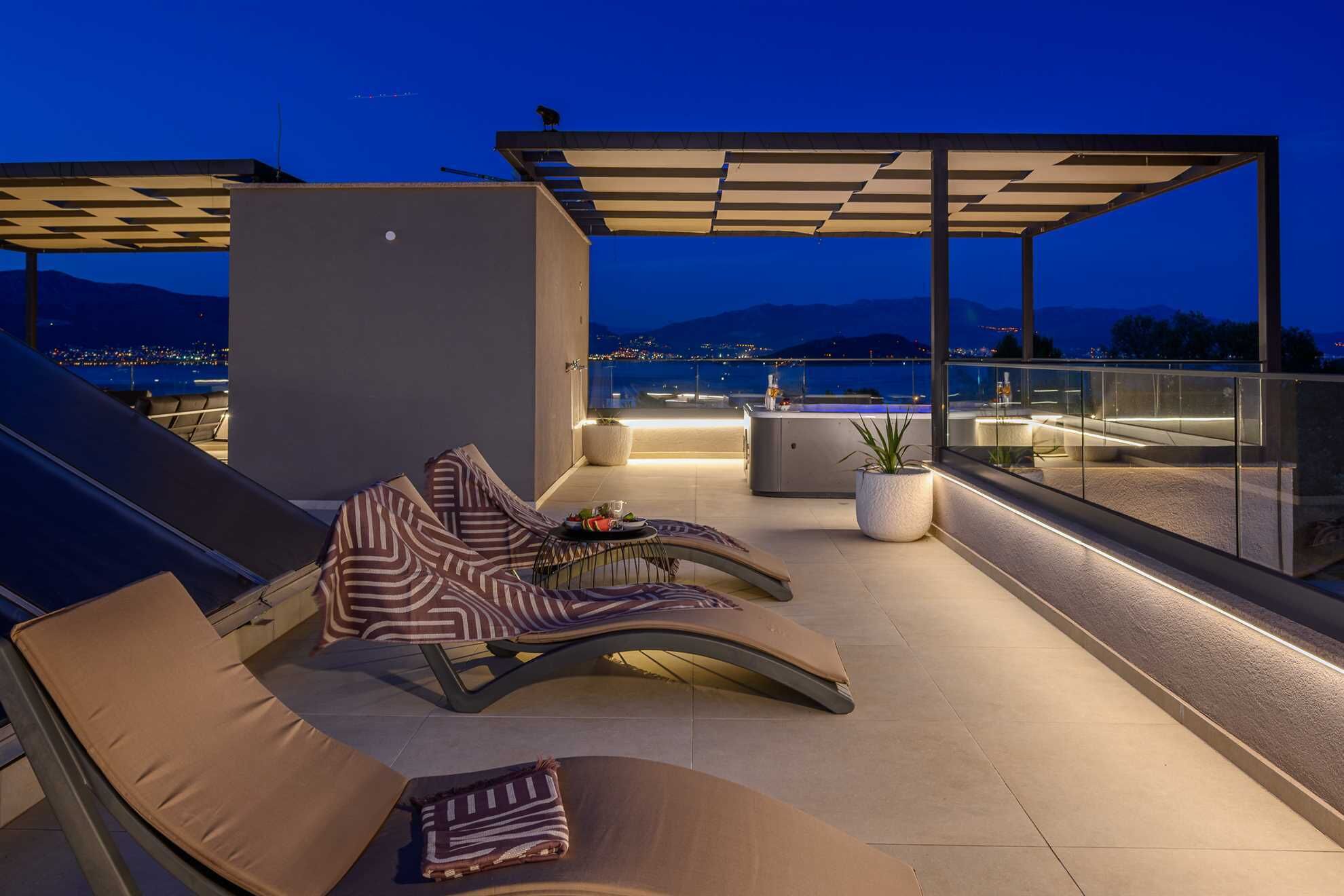 Eine Dachterrasse mit Pool, Liegestühlen und Blick auf die nächtliche Stadt.
