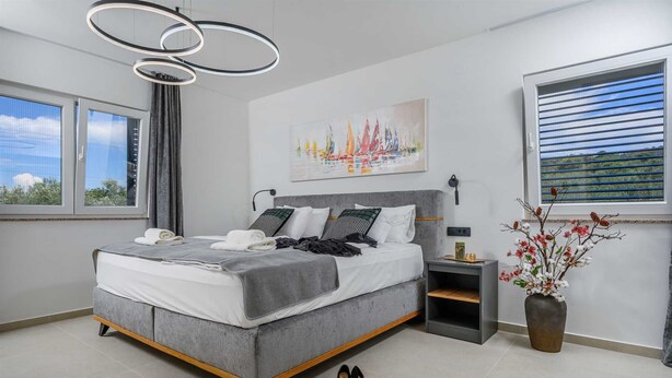 A modern bedroom with a grey upholstered bed, a bedside table, a nightstand, a vase with flowers, and a cityscape painting.