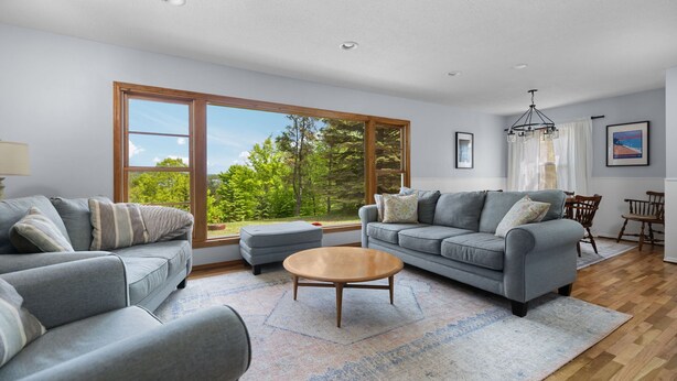 A spacious living room with a large window, two grey sofas, a wooden coffee table, and a dining area with a chandelier. - Woodlea Cottage in the Heart of the National Park (Empire)