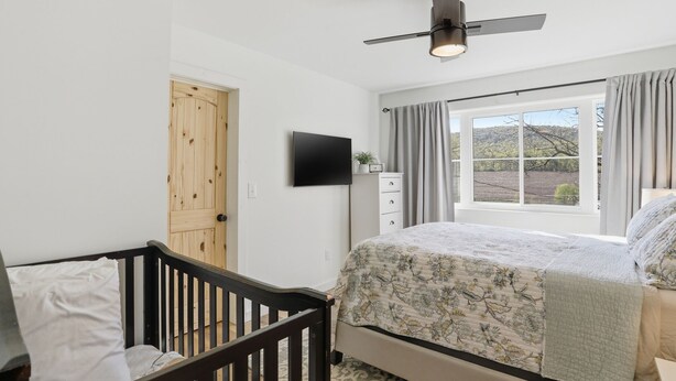 A bedroom with a bed, a crib, a ceiling fan, a TV, a window with curtains, and a view of the outdoors. - Doodle's Mountain Cape - Raystown Lake - Aunt Susie's (James Creek)