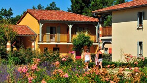 Um edifício de dois andares com telhado de telhas vermelhas, varandas e um jardim com diversas flores.