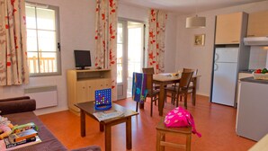 Um quarto com sofá, uma mesa com jogo de tabuleiro, televisão, mesa de jantar com cadeiras, cozinha equipada com frigorífico e janela com cortinas.