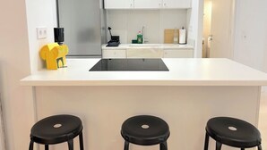 Ett modernt kök med en vit bänkskiva, en inbyggd spis, ett kylskåp och en diskho. Framför bänkskivan står tre svarta barstolar.