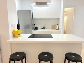 Eine moderne Küche mit weißer Arbeitsplatte, einem integrierten Herd, einem Kühlschrank und einer Spüle. Vor der Arbeitsplatte befinden sich drei schwarze Barhocker.