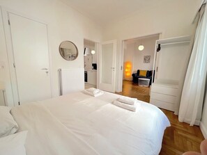 A bedroom with a large bed, a round mirror, a radiator, a kitchen in the background, and a hallway leading to another room. - Modern 2BR Exarchia Apt I Walk to Metro & Museums (Athina)