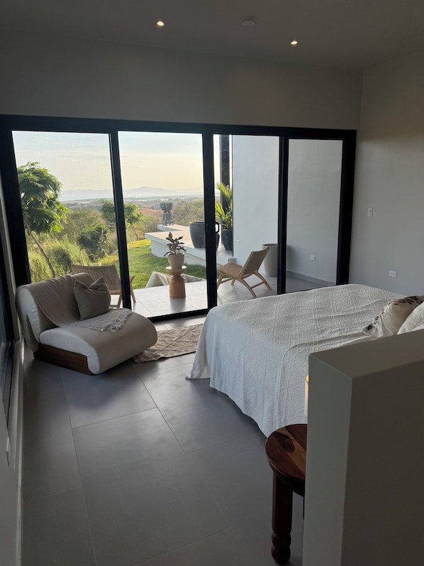 A bedroom with a large sliding glass door leading to a balcony, a bed, a chair, and a small table.