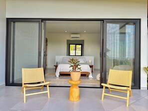 A bedroom with a bed, two wicker chairs, a wooden table, and a sliding glass door leading to a balcony.