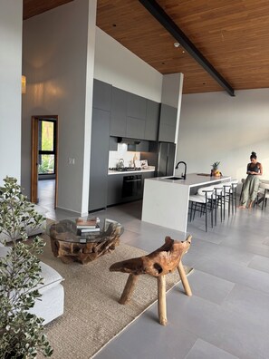 A modern kitchen with a central island, a dining area with bar stools, and a living space with a glass coffee table and wooden side table.