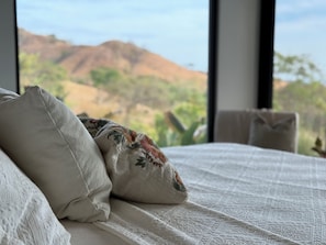 A bed with pillows, a window with a view of a mountain and greenery.