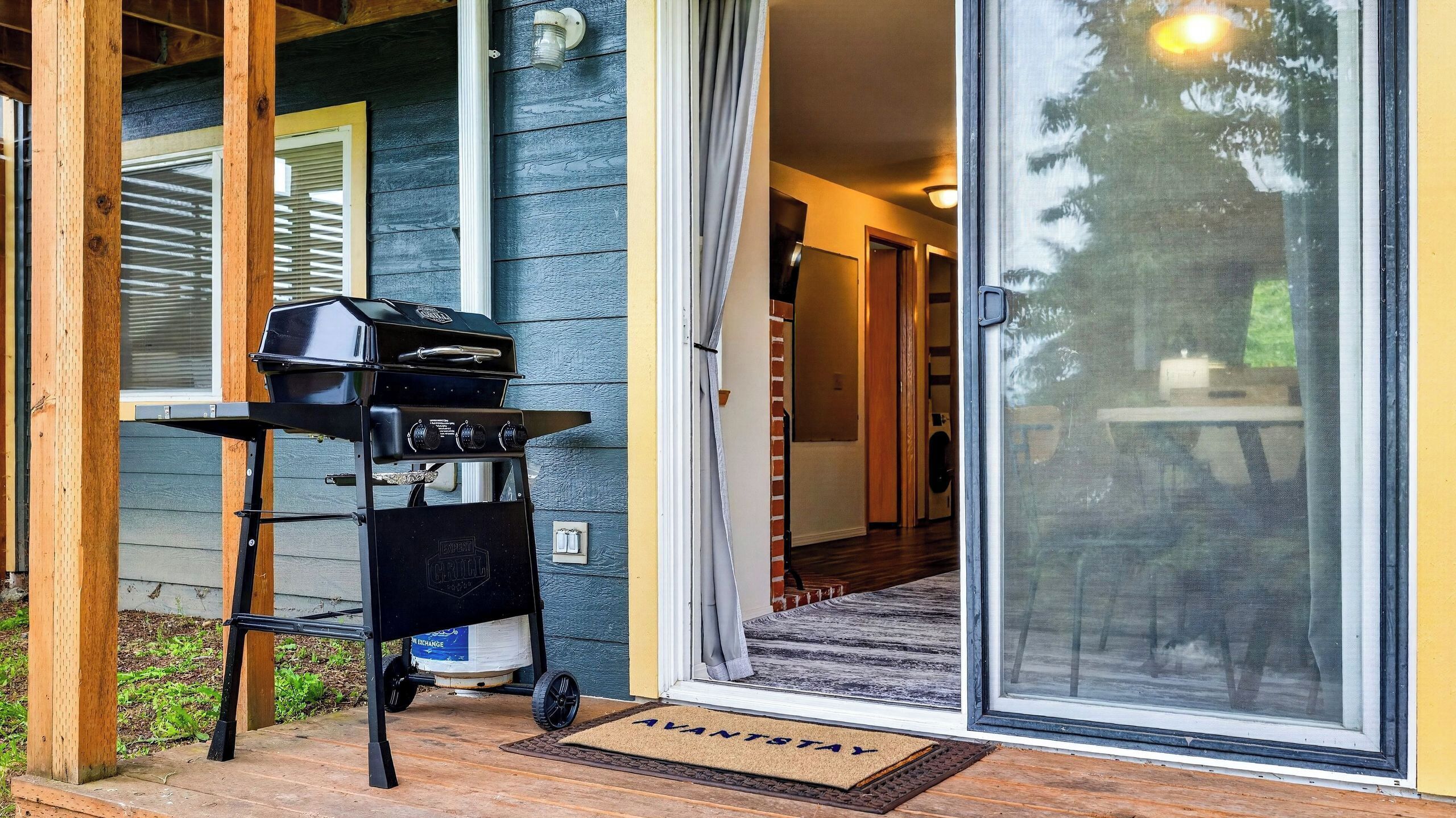Una casa con puerta corrediza de vidrio que da a un porche cubierto, una parrilla de barbacoa y un felpudo con la inscripción 'AVANTSTAY'.