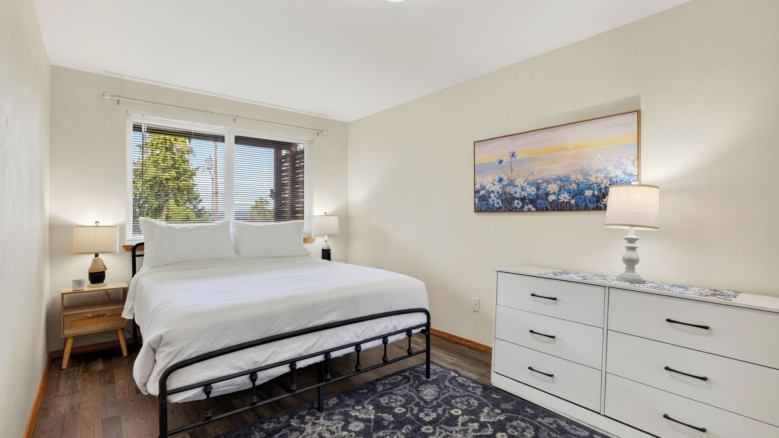 Een slaapkamer met een bed, nachtkastjes, een dressoir, een raam met zonwering en een schilderij aan de muur.