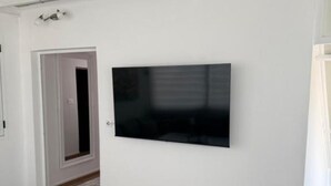 Huoneessa on seinälle asennettu litteänäyttöinen televisio, ovi, joka johtaa toiseen huoneeseen, ja laatoitettu lattia.