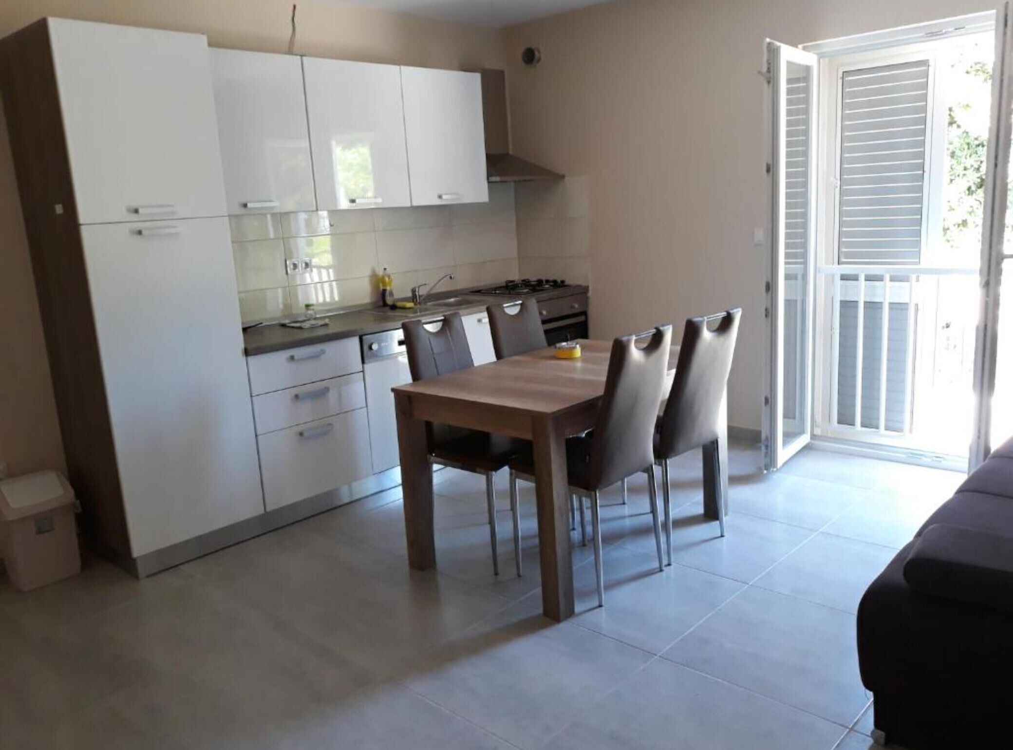 Une cuisine moderne avec des meubles blancs, une table à manger avec des chaises et une porte-fenêtre coulissante donnant sur un balcon.