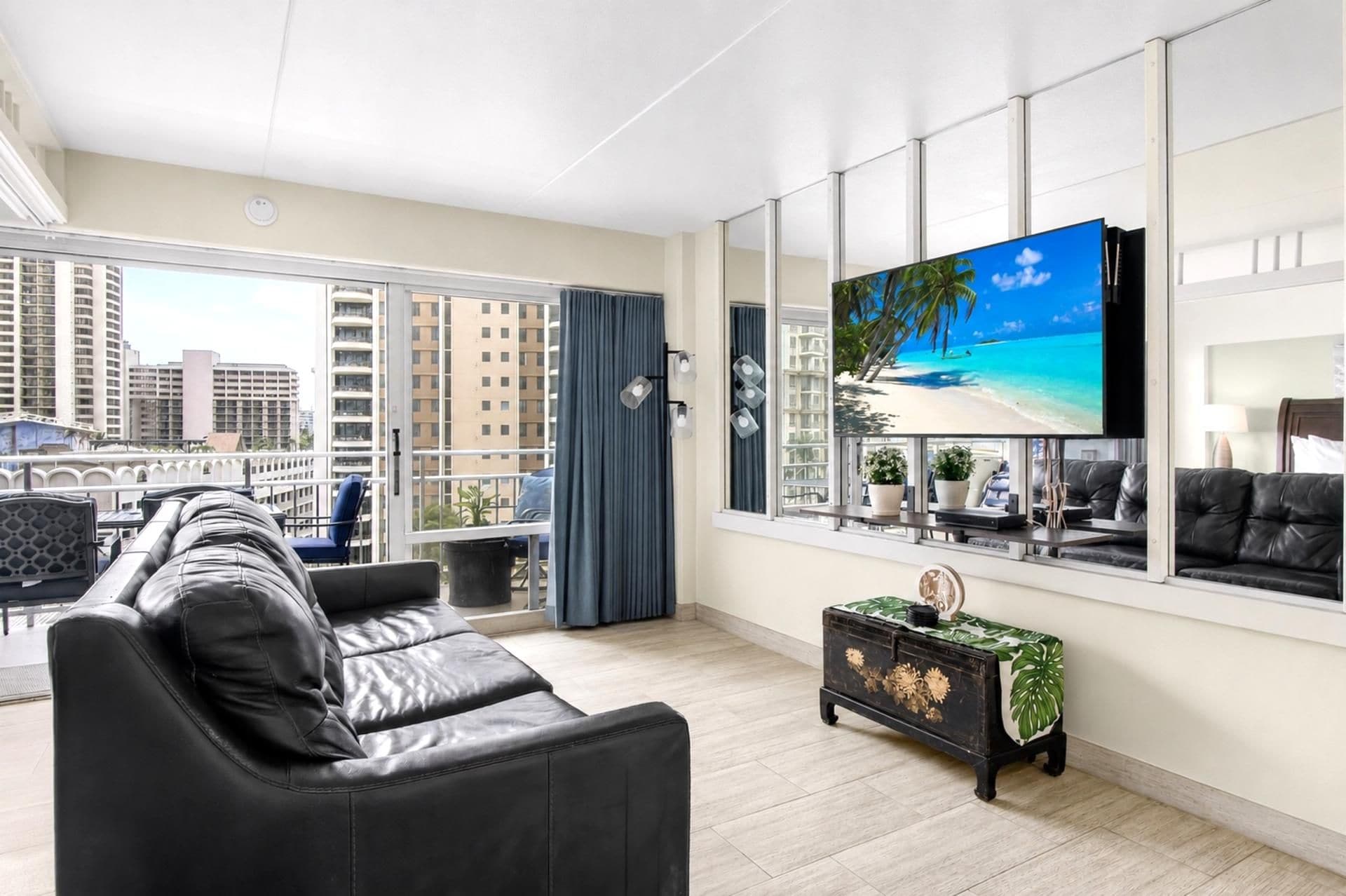 A modern living room with a black leather sofa, a small side table with a decorative item, a large TV screen displaying a beach scene, and a balcony with a view of tall buildings.