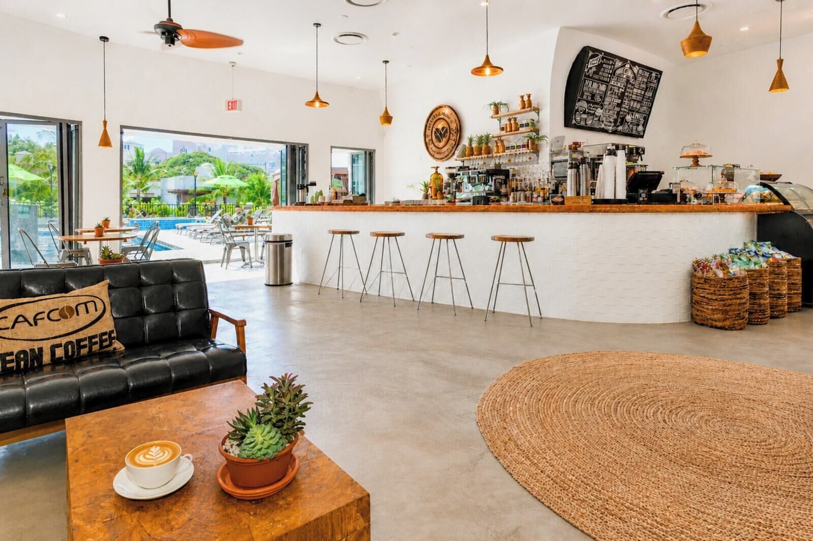 Interior moderno de una cafetería con barra, zona de estar y amplios ventanales con vistas a una piscina.