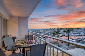 Ein Balkon mit Essplatz, der auf einen Hafen mit Booten und eine städtische Skyline bei Sonnenuntergang blickt.