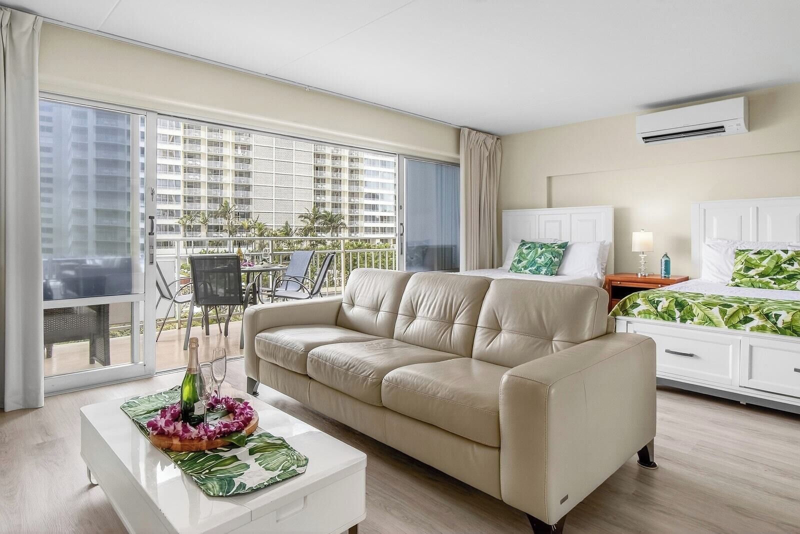 A modern hotel room with a beige sofa, a white bedside table, a bed with a green leaf-patterned bedspread, and a balcony with outdoor furniture.