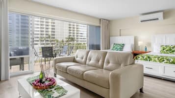 A modern hotel room with a beige sofa, a white bedside table, a bed with a green leaf-patterned bedspread, and a balcony with outdoor furniture.