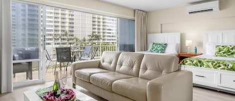 Une chambre d’hôtel moderne avec un canapé beige, une table de chevet blanche, un lit recouvert d’une couvre-lit à motifs de feuilles vertes et un balcon aménagé avec du mobilier d’extérieur.