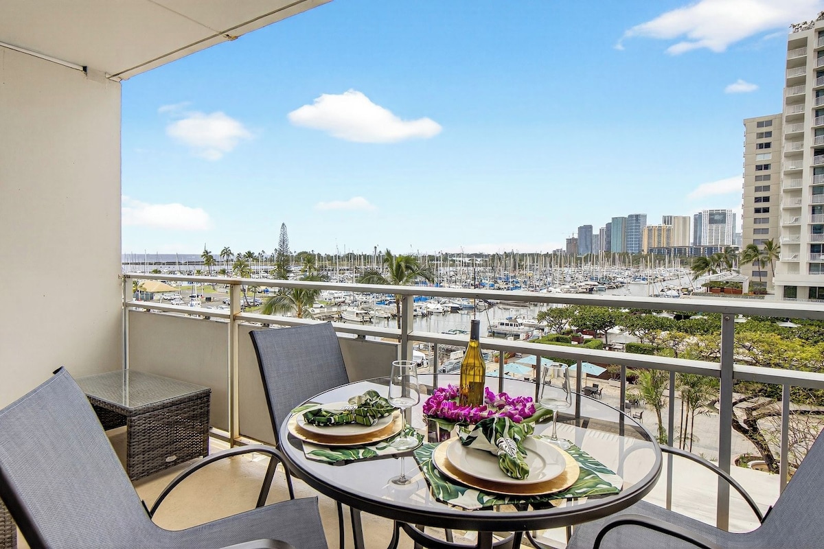 Un balcón con una mesa dispuesta para dos personas, con vistas a un puerto deportivo con barcos y edificios de la ciudad.