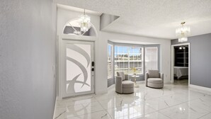 Una habitación amplia con piso de mármol, una puerta blanca con diseño geométrico, dos sillones, una mesa pequeña con un jarrón y un candelabro.