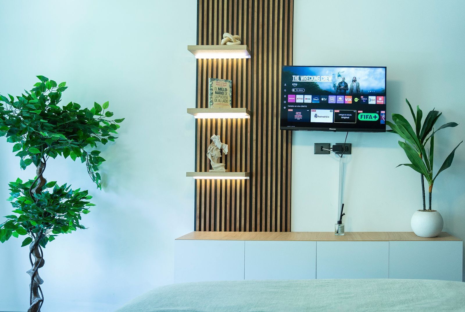 Et moderne værelse med et TV ophængt på væggen, en træhylde med dekorative genstande og en potteplante.