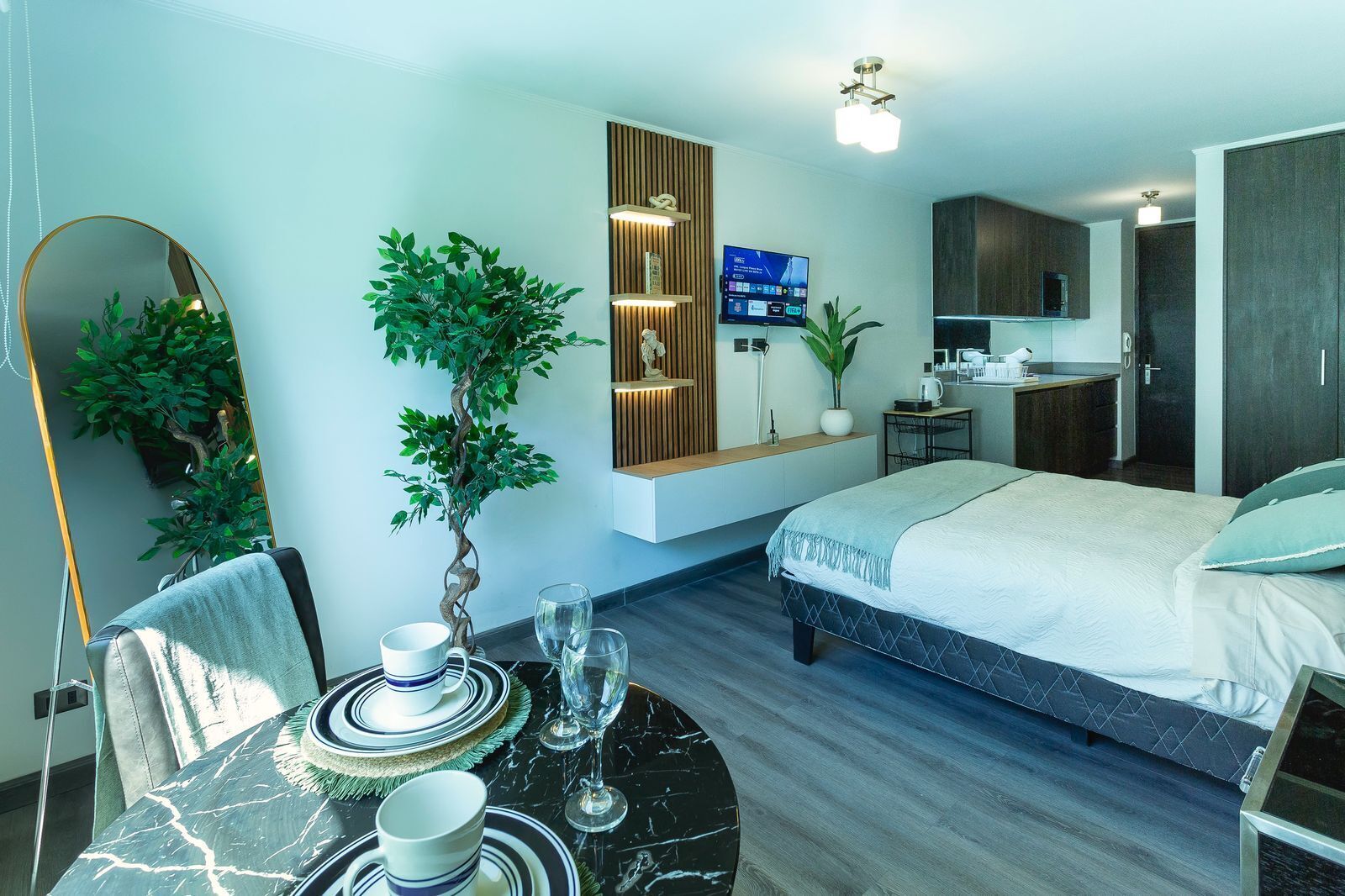 Ein modernes Hotelzimmer mit einem Bett, einem Esstisch mit Tellern und Gläsern, einem Fernseher, einer kleinen Küchenzeile und einer Topfpflanze.