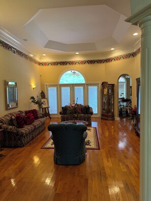 A spacious living room with hardwood flooring, a large arched window, and a grand piano. - AU GAMEDAY Private Lake House, Fishing (Auburn)