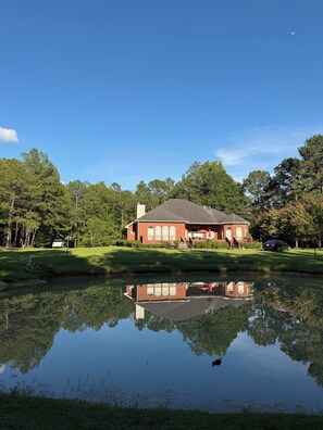 A red brick house with a reflection in a pond, surrounded by trees and a clear sky. - AU GAMEDAY Private Lake House, Fishing (Auburn)