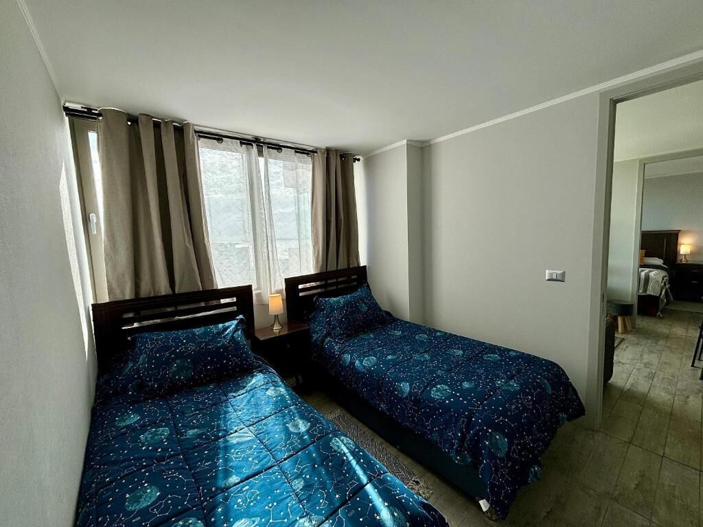 A room with two beds, a window with curtains, a nightstand, and a visible bathroom area.