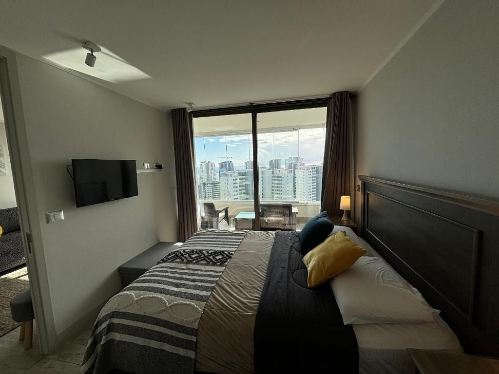 A hotel room with a bed, a television, a balcony with a city view, and a window with curtains.