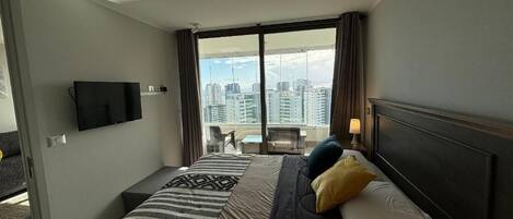 Habitación de hotel con cama, televisor, balcón y vistas a la ciudad, y ventana con cortinas.