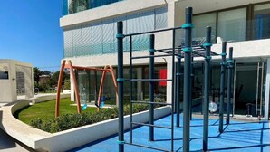 Zona de juegos al aire libre con estructura de escalada y columpios, junto a un edificio con amplios ventanales.