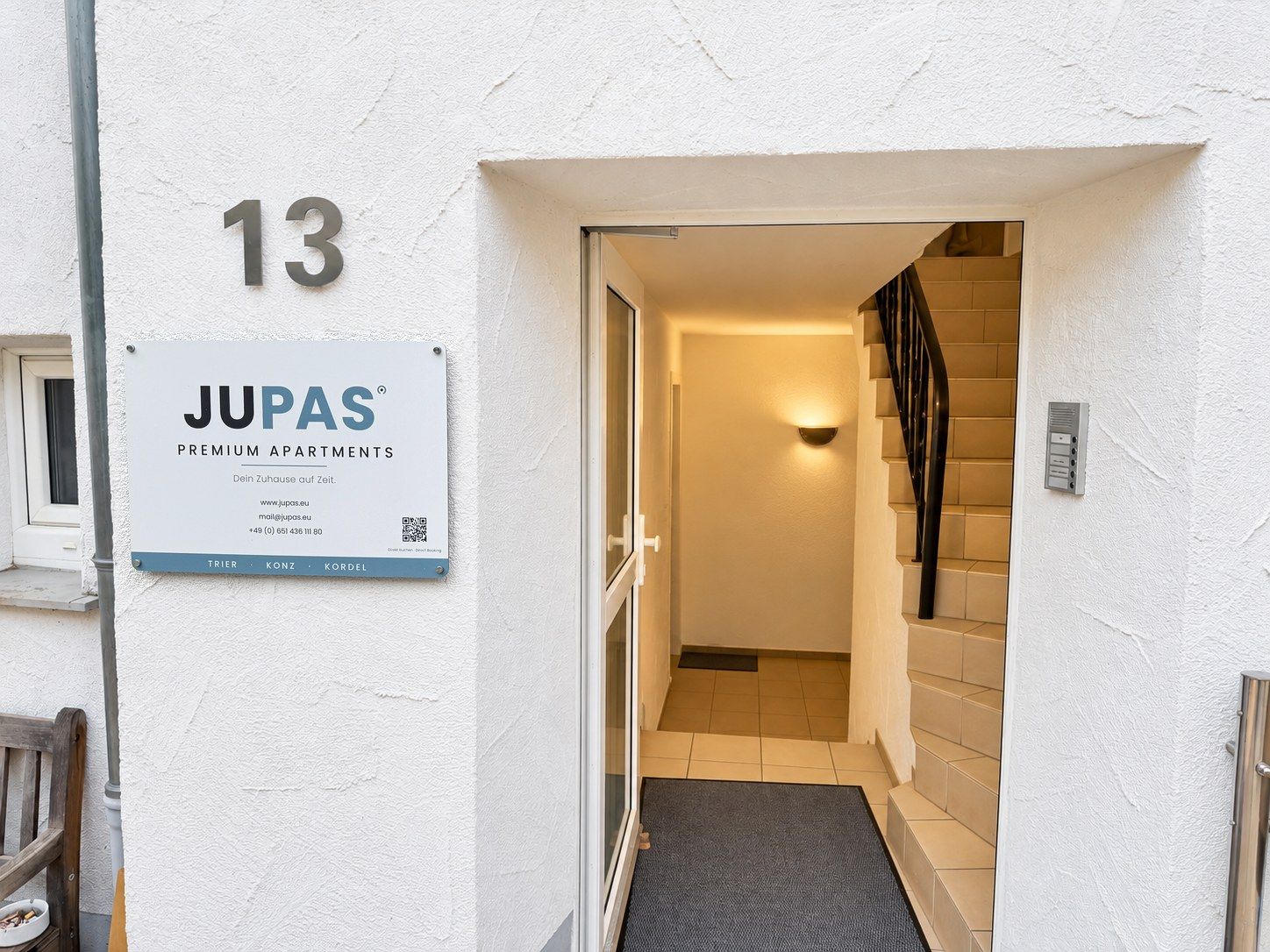 Une entrée d’immeuble d’habitation avec une plaque portant le numéro 13, une enseigne « JUPAS » et un escalier menant à un appartement.