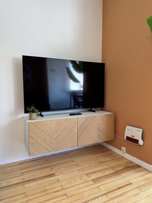 A modern living room with a flat-screen TV mounted on a wooden cabinet, a small potted plant, and a wall-mounted thermostat. - JUPAS° | Saar Home - Nature & Relaxed Vibes (Konz)