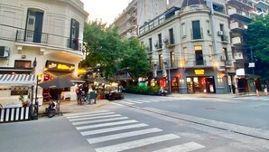 Una esquina de calle con edificios, un paso de peatones y mesas al aire libre en un café.