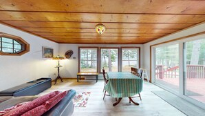 A cozy room with a wooden ceiling, a table with a striped tablecloth, a bench, a lamp, and large windows overlooking a deck. - Pioneer Paradise - Lakefront home (Conover)