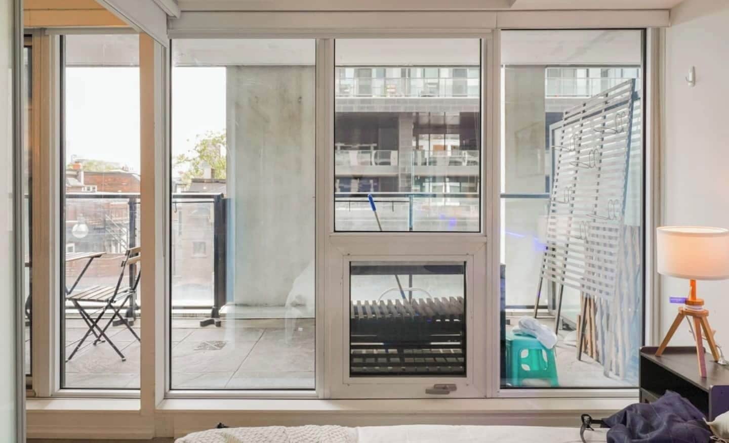 A room with large windows, a view of a cityscape, a radiator, and a lamp on a side table.