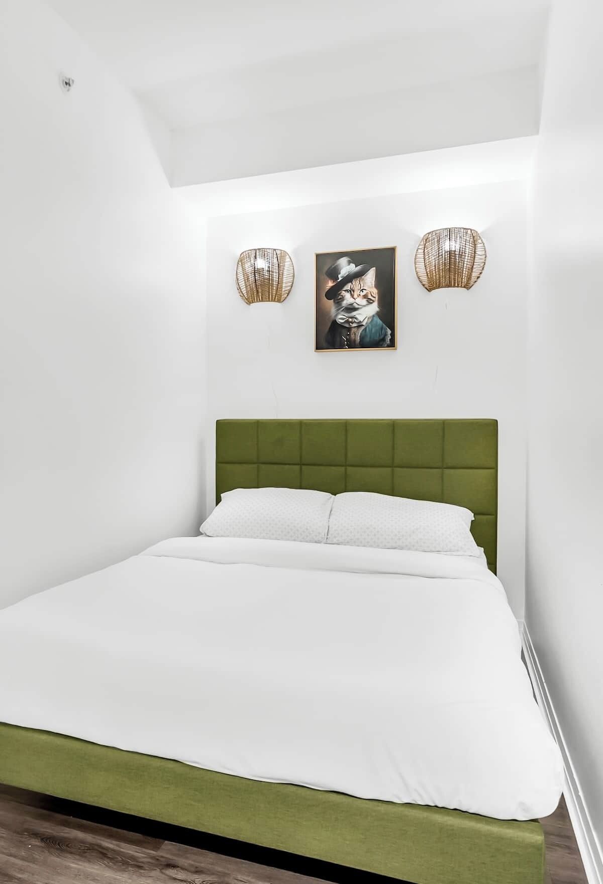 Une chambre à coucher avec une tête de lit recouverte d’un tissu vert, une couvre-lit blanche et deux lampes fixées au mur.
