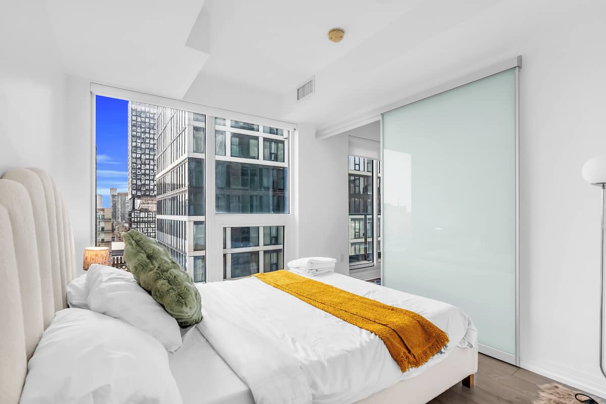 A modern hotel room with a large bed, white bedding, a mustard-colored throw blanket, and a view of a cityscape through floor-to-ceiling windows.