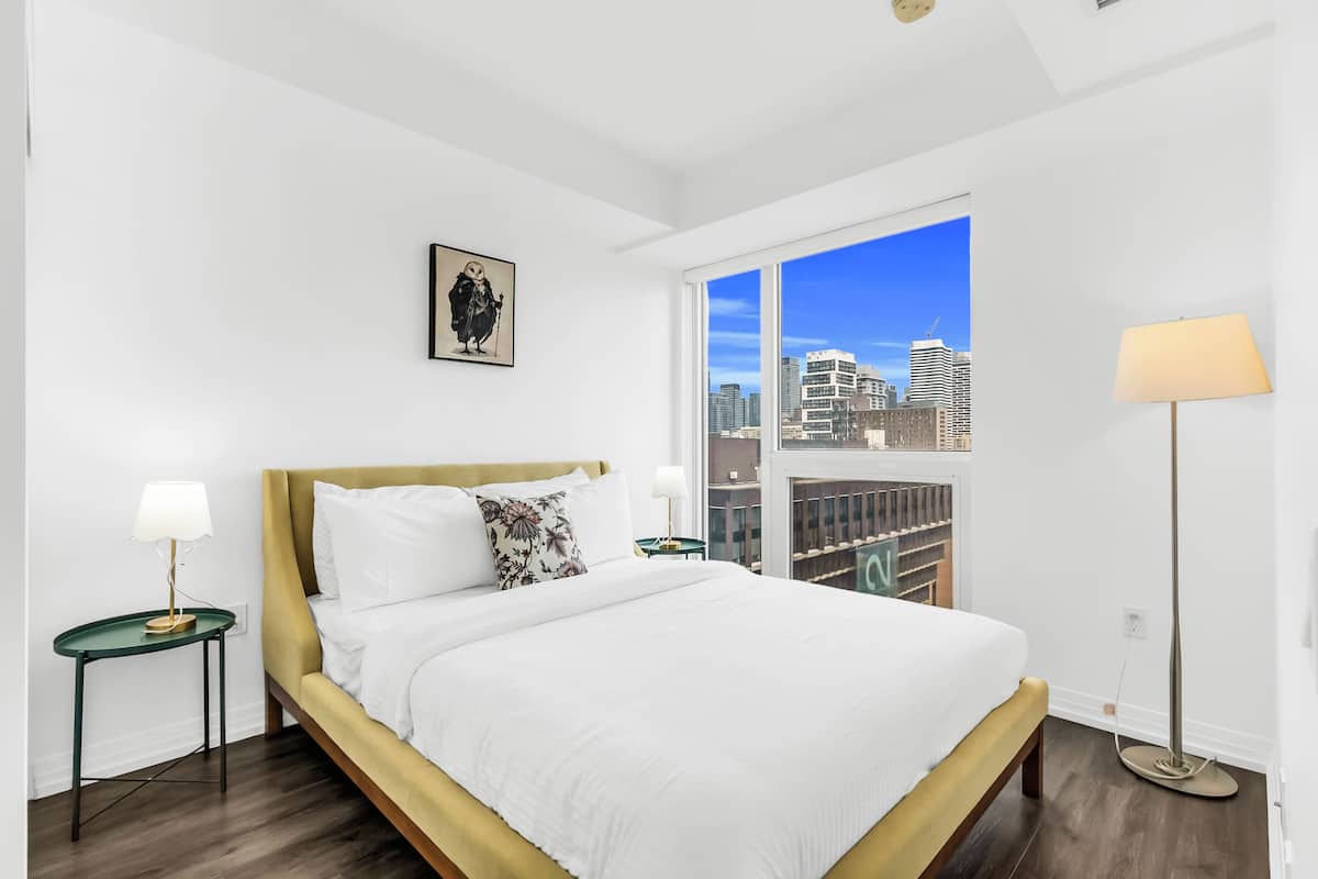 A modern bedroom with a large bed, a city view, and a framed artwork on the wall.