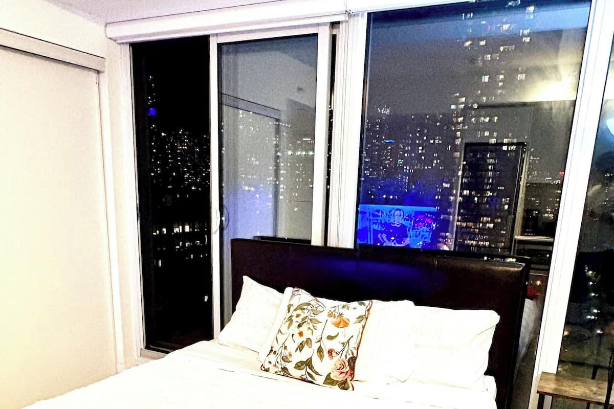 A hotel room with a large bed, a red throw blanket, a small plant, and a city view through large windows.
