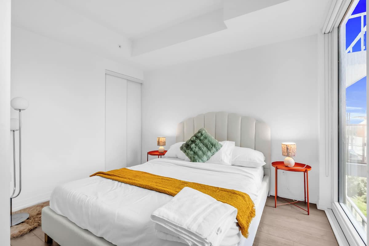 A modern bedroom with a large bed, white bedding, a mustard throw blanket, and two bedside tables with lamps.