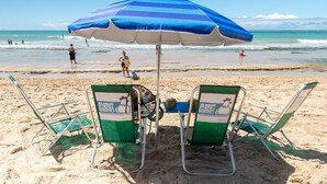 Ein Strand mit einem blau-weiß gestreiften Sonnenschirm, zwei grünen Liegestühlen und einer Kokosnuss im Sand.