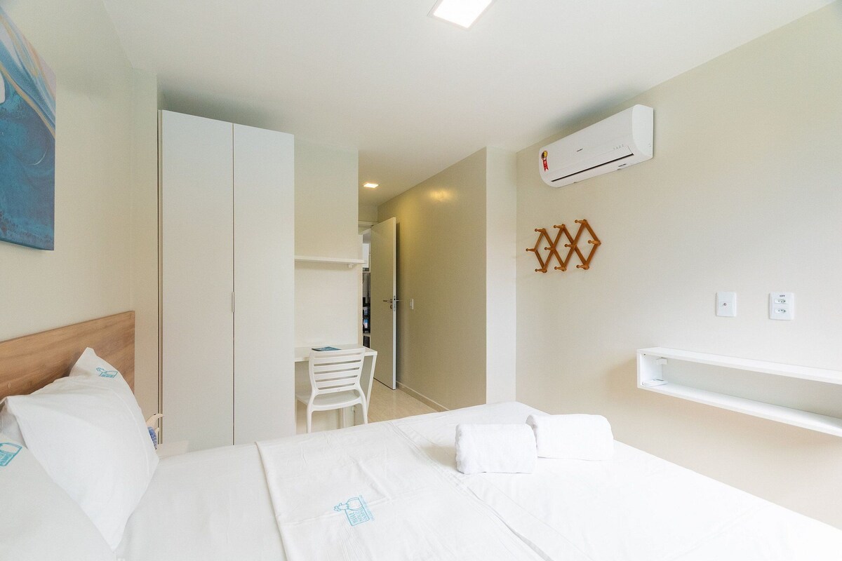 A hotel room with a bed, a desk, a chair, an air conditioning unit, and a wall-mounted shelf.