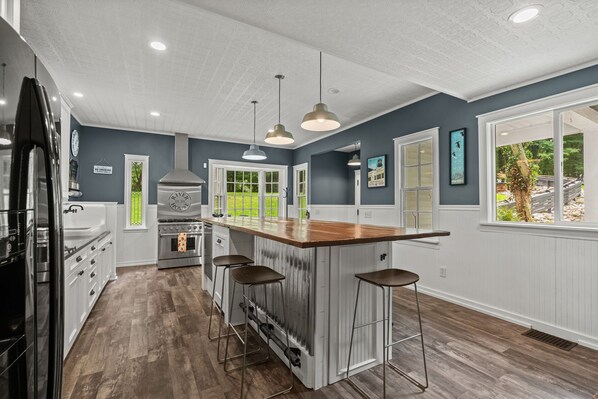 A kitchen with a central island, bar stools, a stainless steel range hood, and a view of the outdoors through a window. - River House in Fennville (Fennville)