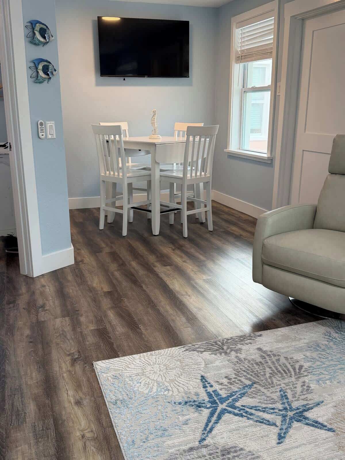 Un coin repas avec une table et des chaises, un téléviseur fixé au mur et une fenêtre avec des stores.
