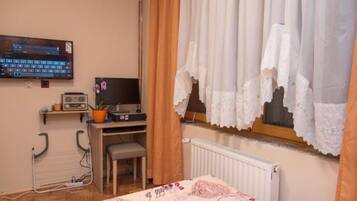 A bedroom with a bed, a desk with a computer, a chair, a radiator, and a window with curtains.