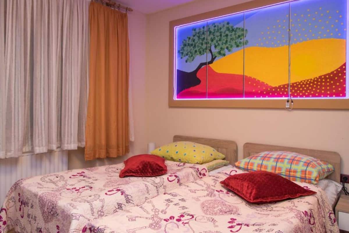 Un dormitorio con cama, una ventana y cortinas, junto a un cuadro de colores.