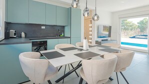 A modern kitchen with teal cabinets, a dining area with a table and chairs, and a large window with a view of a boat. - Villa Pomalo (Vodice)